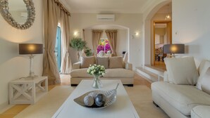 A living room with a sofa, a coffee table with decorative items, a lamp, and a mirror. There is a view of a kitchen and a dining area in the background.