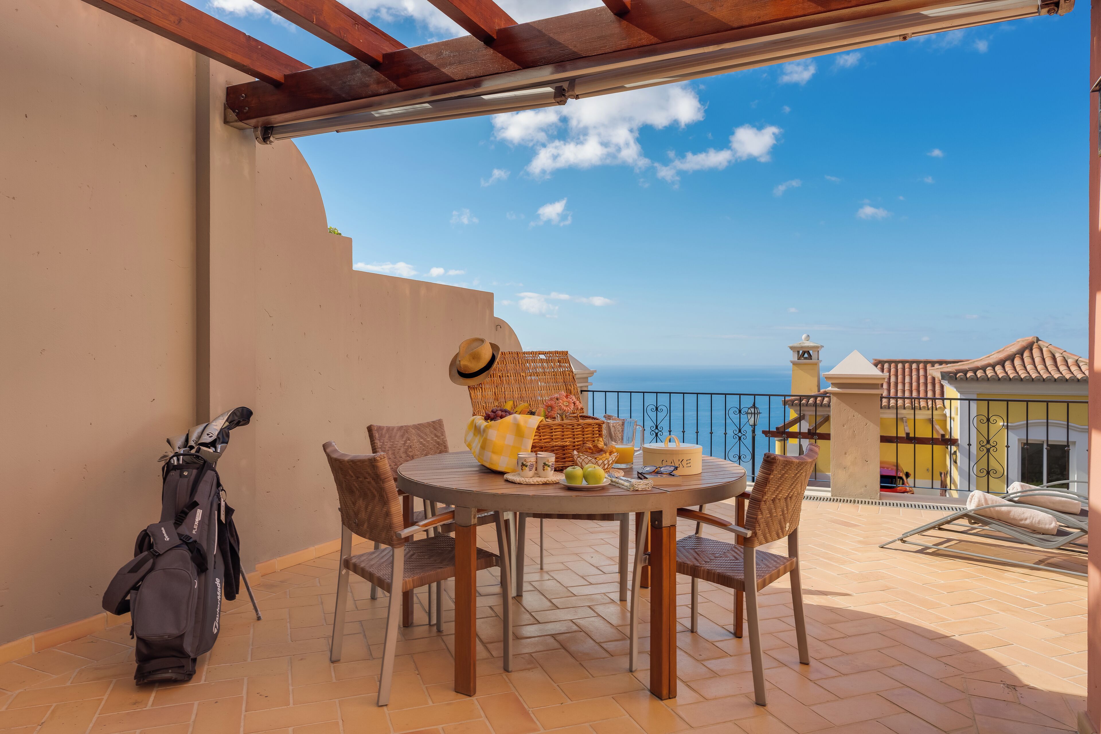 A round table with chairs, a wicker basket, and a golf bag on a balcony with a view of the sea.