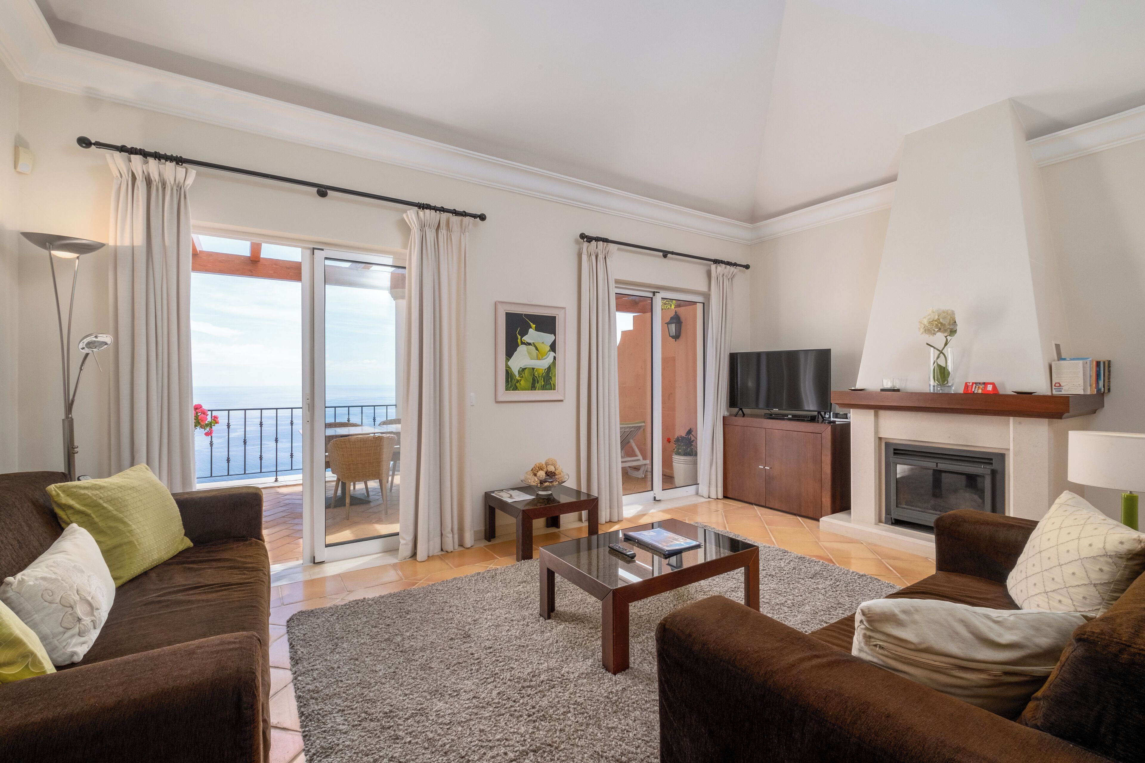 Een woonkamer met een bank, salontafel en open haard. Er is een balkon met gordijnen en uitzicht op zee.
