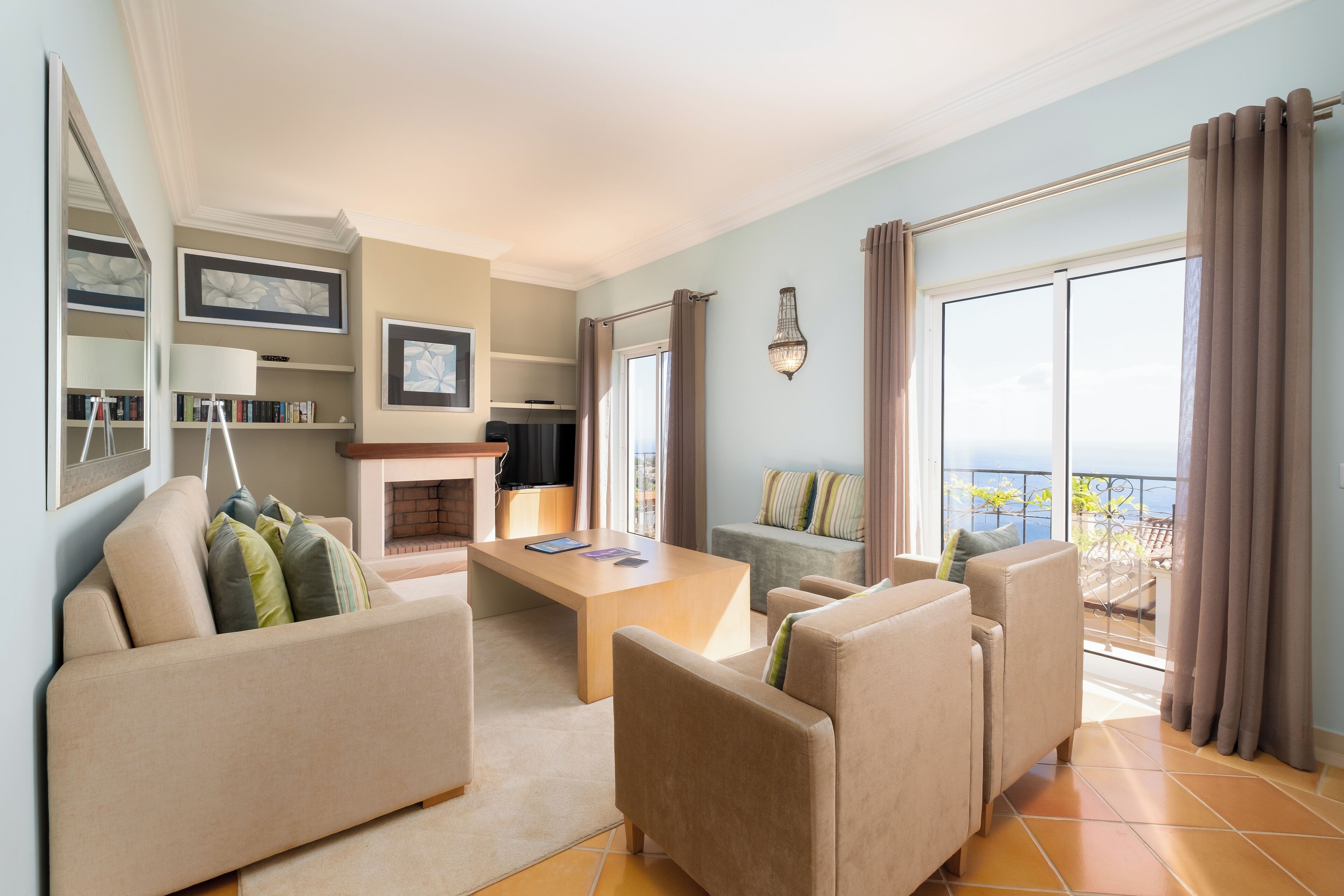 Een moderne woonkamer met een bank, fauteuils, een salontafel en een open haard. Er hangen ingelijste foto’s aan de muur en er is een groot raam met gordijnen.