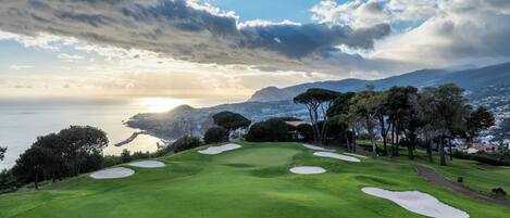 A golf course with sand bunkers, a pathway, and a view of the sea and mountains in the background.