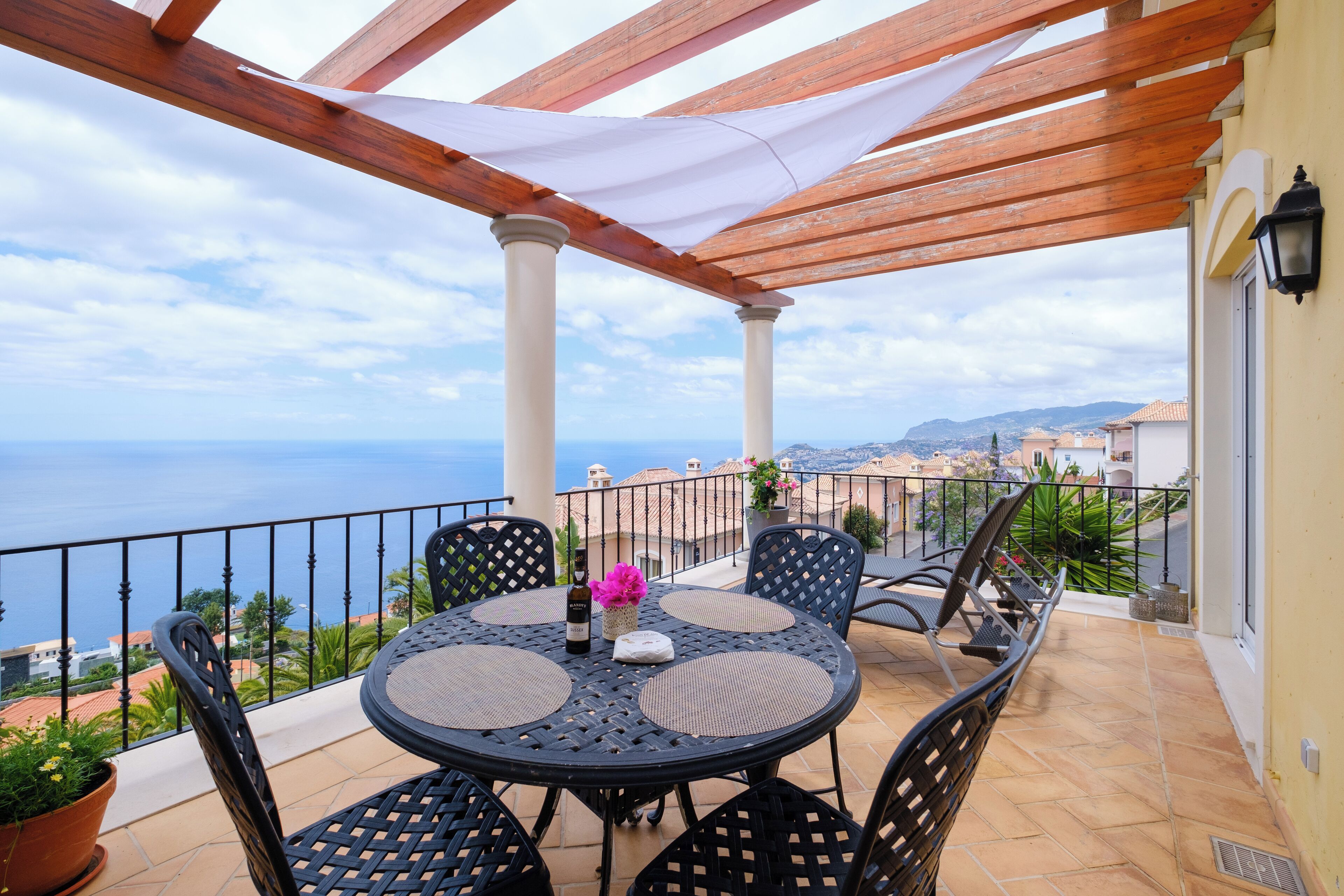 A covered outdoor seating area with a view of the sea, featuring a round table with chairs and a potted plant.