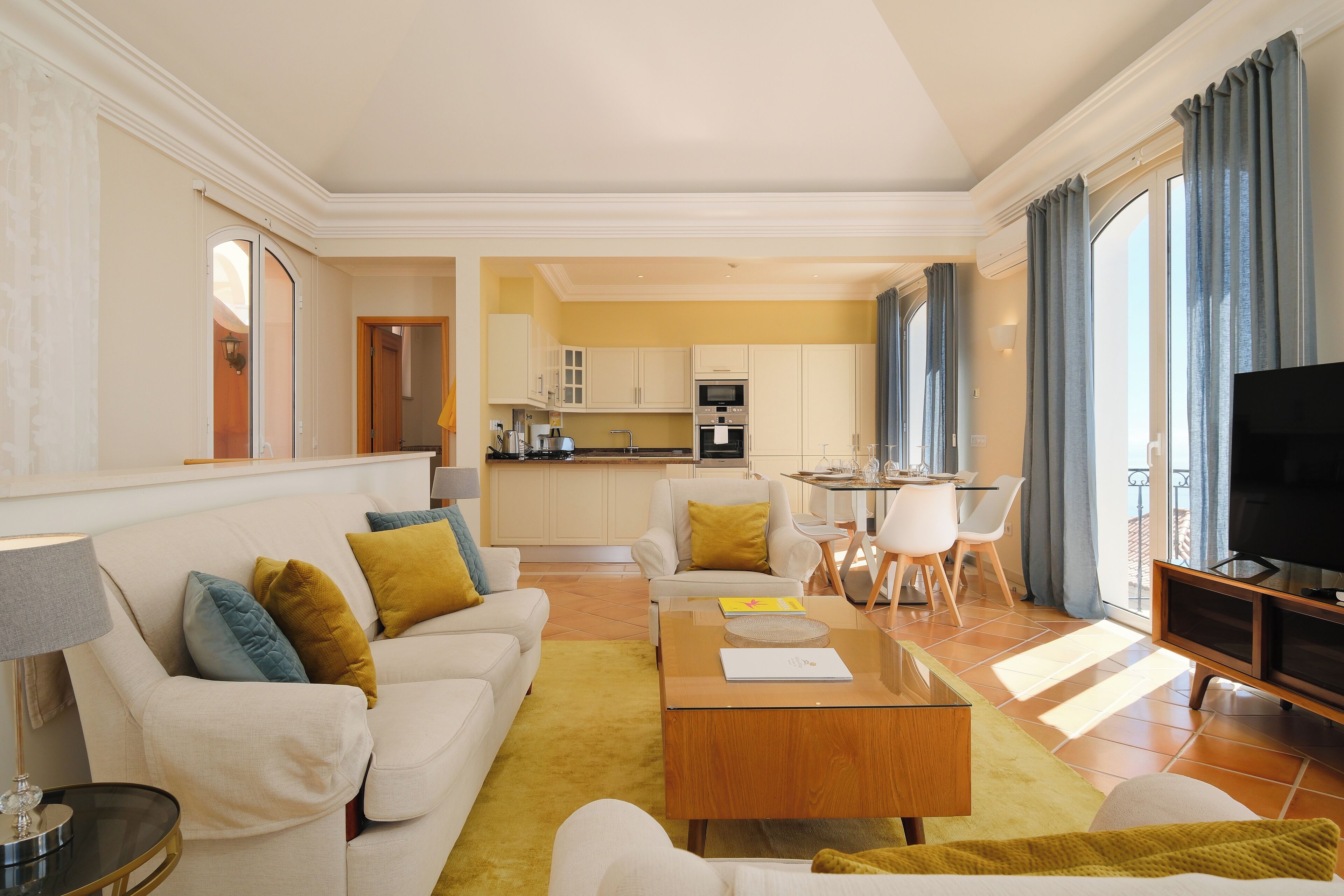 A modern living room with a sofa, coffee table, and a TV. There is a kitchen in the background with a dining area.