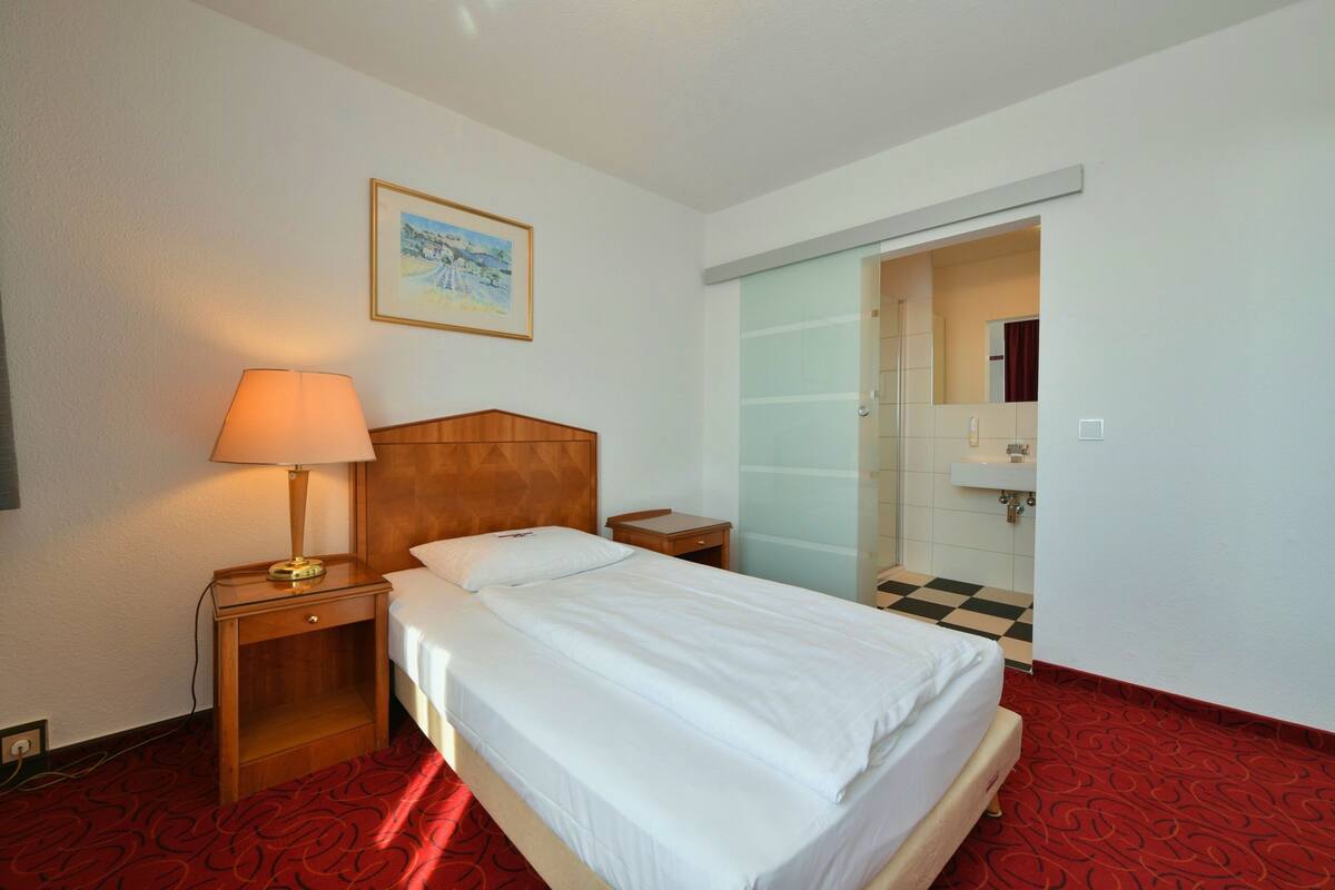 A hotel room with a bed, a nightstand with a lamp, a wooden headboard, a framed picture on the wall, and a bathroom visible through a sliding door.