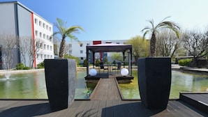 A wooden deck leading to a covered seating area with a pond, surrounded by buildings and palm trees. - SKYHOTEL Merseburg (Merseburg)