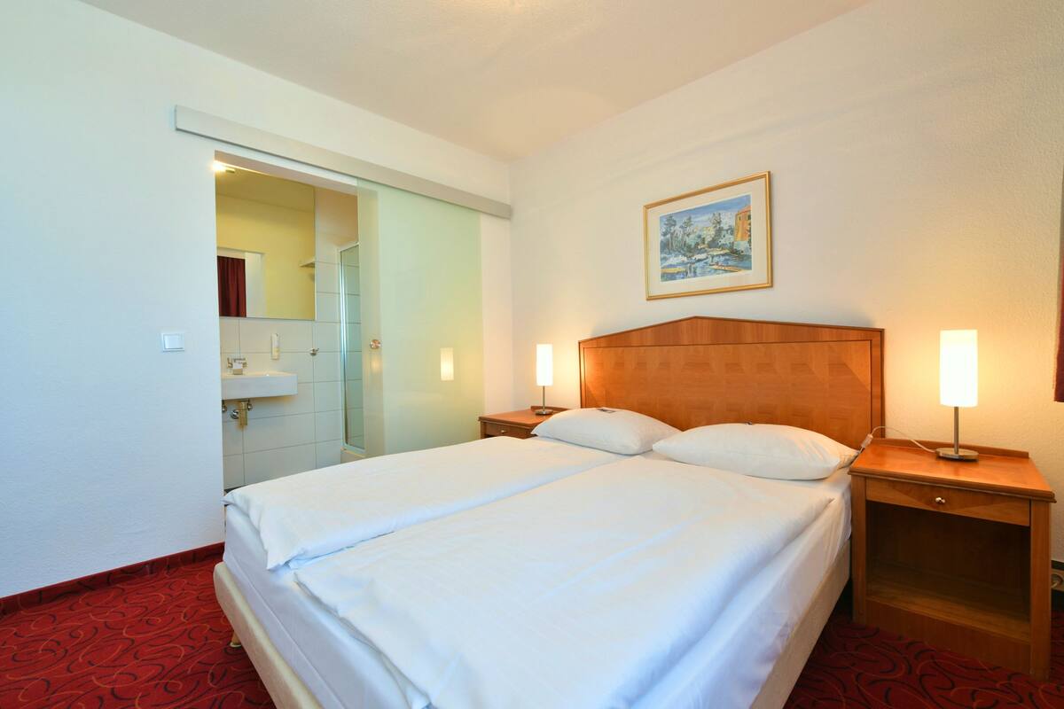 A hotel room with a large bed, two bedside tables with lamps, a painting on the wall, and a bathroom visible through an open door.