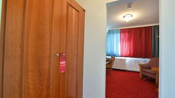 Ein Hotelzimmer mit einem Holfischschrank, einem roten Teppich, einem Esstisch mit Stühlen und einem Fenster mit roten Vorhängen.