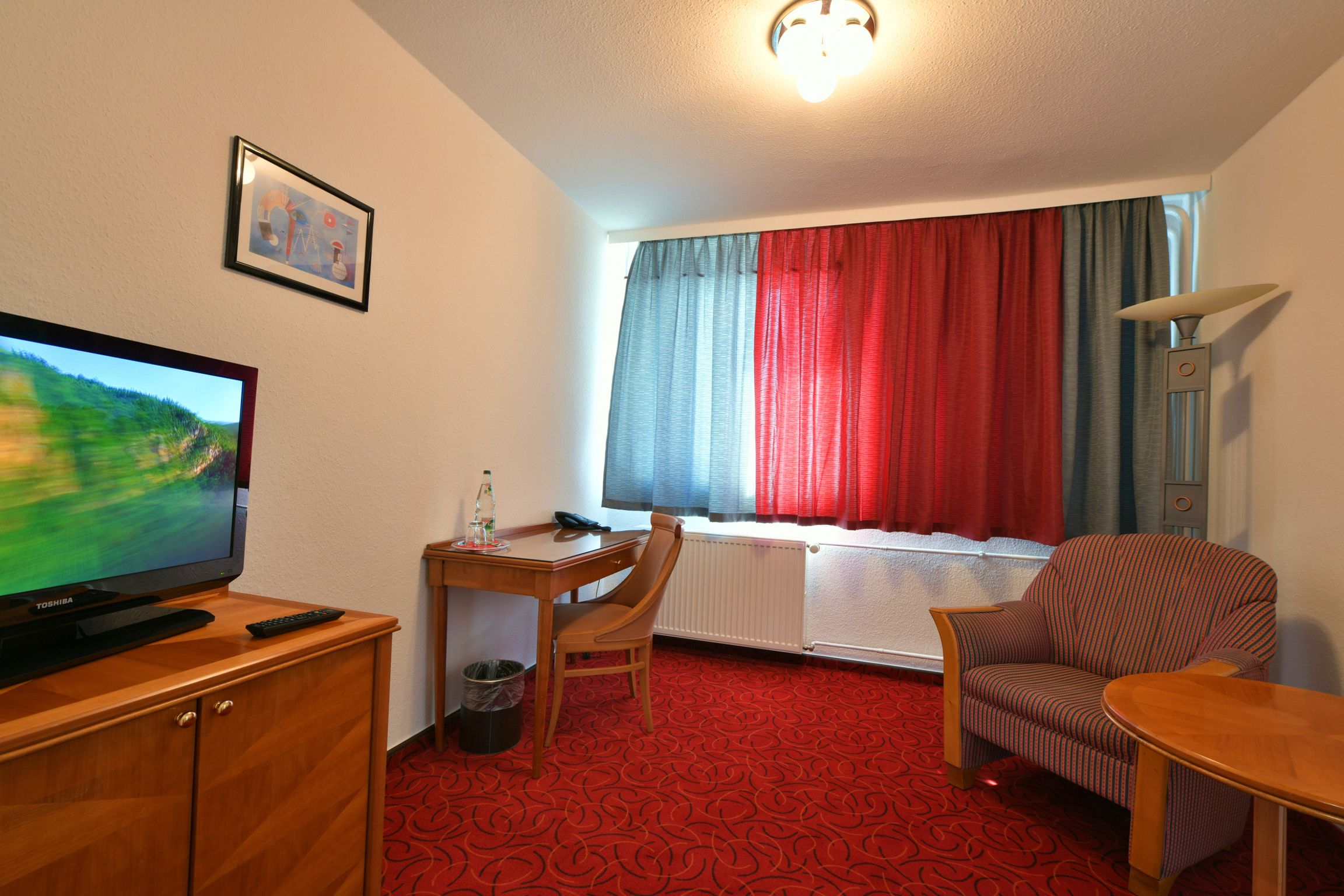 Habitación de hotel con televisión, escritorio con silla, radiador y ventana con cortinas rojas.