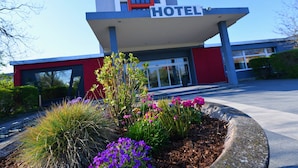 A modern hotel with a red and white facade, a covered entrance, and a landscaped garden with various plants and flowers. - SKYHOTEL Merseburg (Merseburg)
