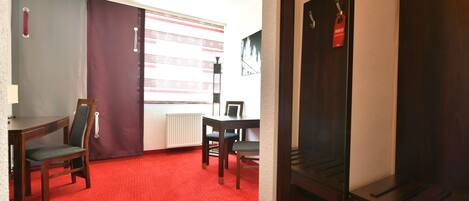 A hotel room with a red carpet, a wooden desk with a chair, a radiator, and a window with curtains.