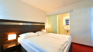 A hotel room with a large bed, a nightstand with a lamp, a red carpet, and a door leading to a bathroom.