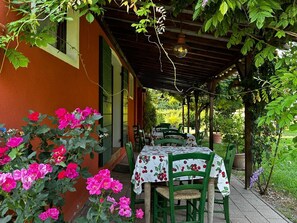 Un área de estar al aire libre cubierta, con una mesa y sillas, rodeada de follaje y plantas con flores.