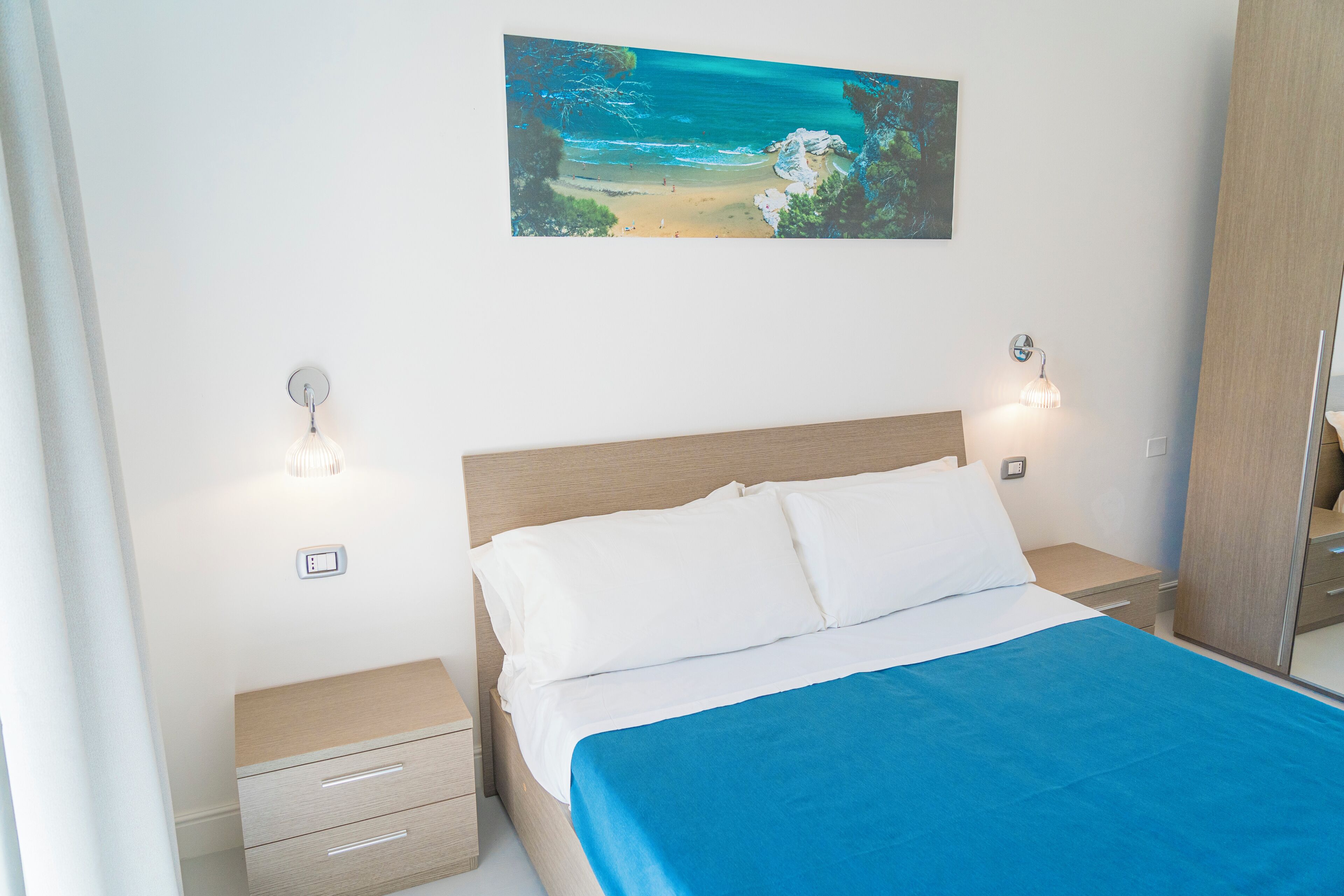 Um quarto com uma cama, mesa de cabeceira, gaveta, luminária na parede e um quadro com uma cena de praia.