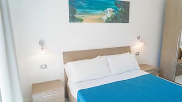 Um quarto com uma cama, mesa de cabeceira, gaveta, luminária na parede e um quadro com uma cena de praia.
