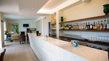 Uma área de bar moderna com um balcão comprido, prateleiras com garrafas e copos, e um espaço de estar com cadeiras e um sofá.