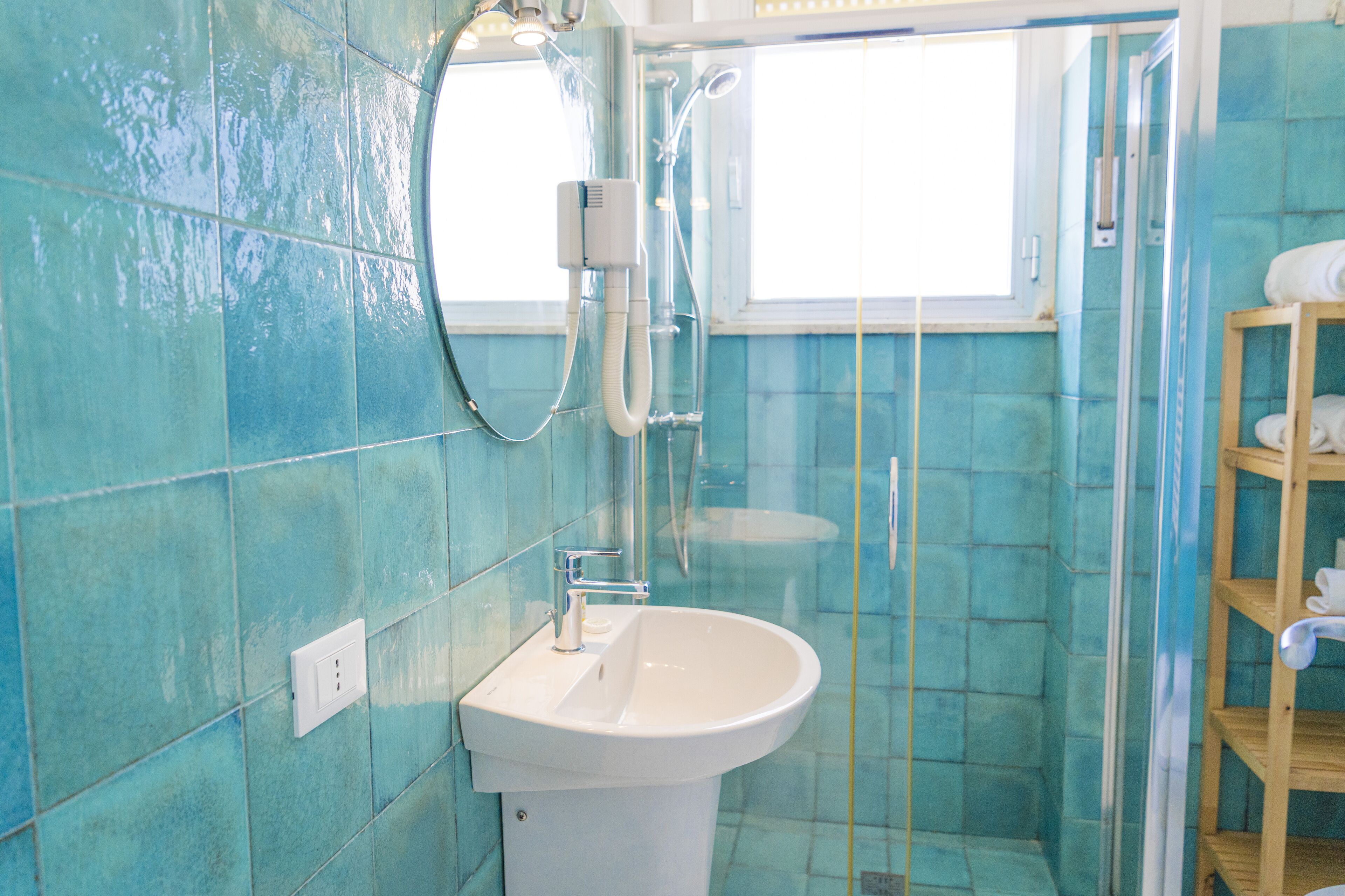 Une salle de bain avec un lavabo blanc, une cabine de douche en verre et une étagère en bois où sont posées des serviettes.