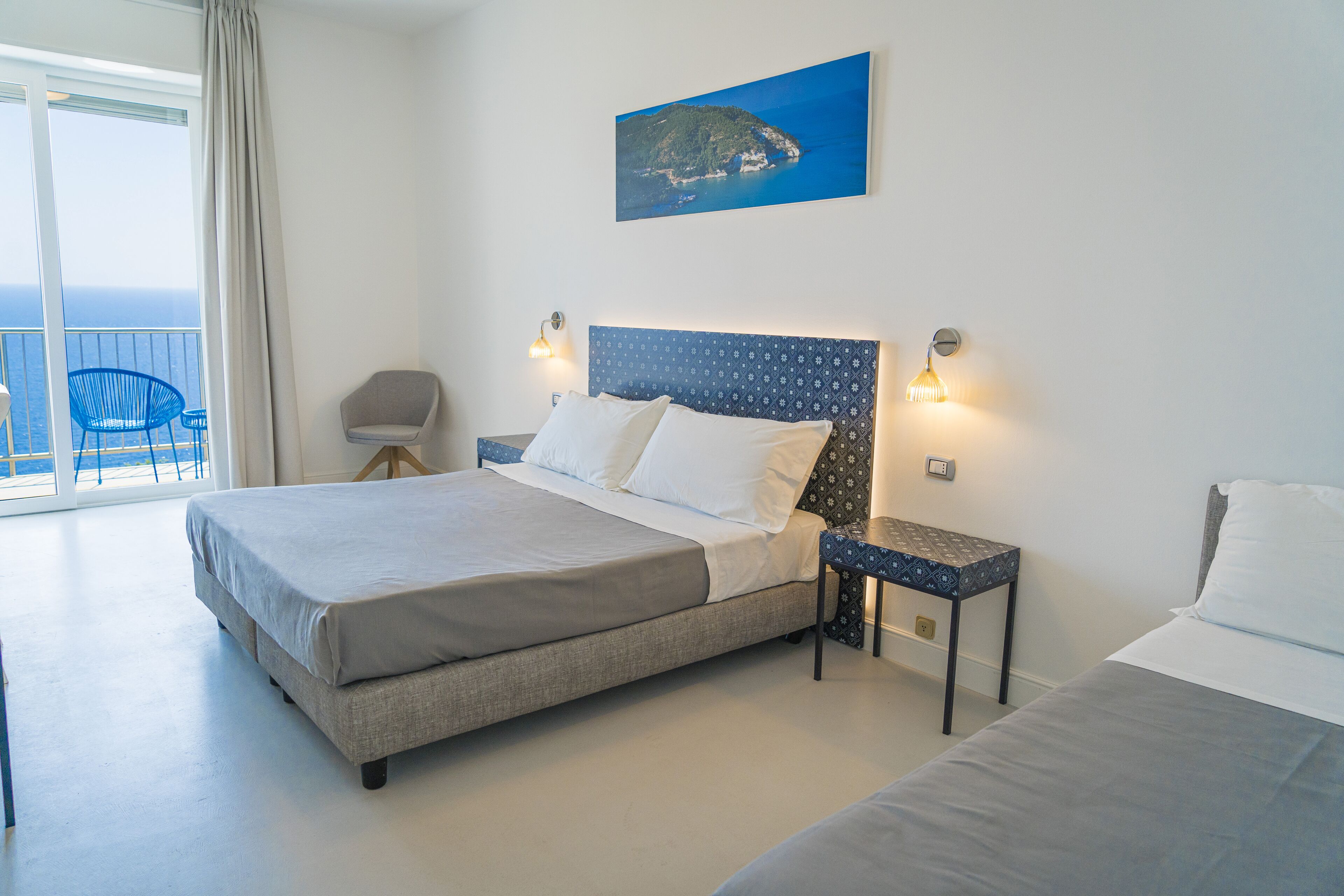 A bedroom with a large bed, a smaller bed, a nightstand, a chair, and a view of the ocean.