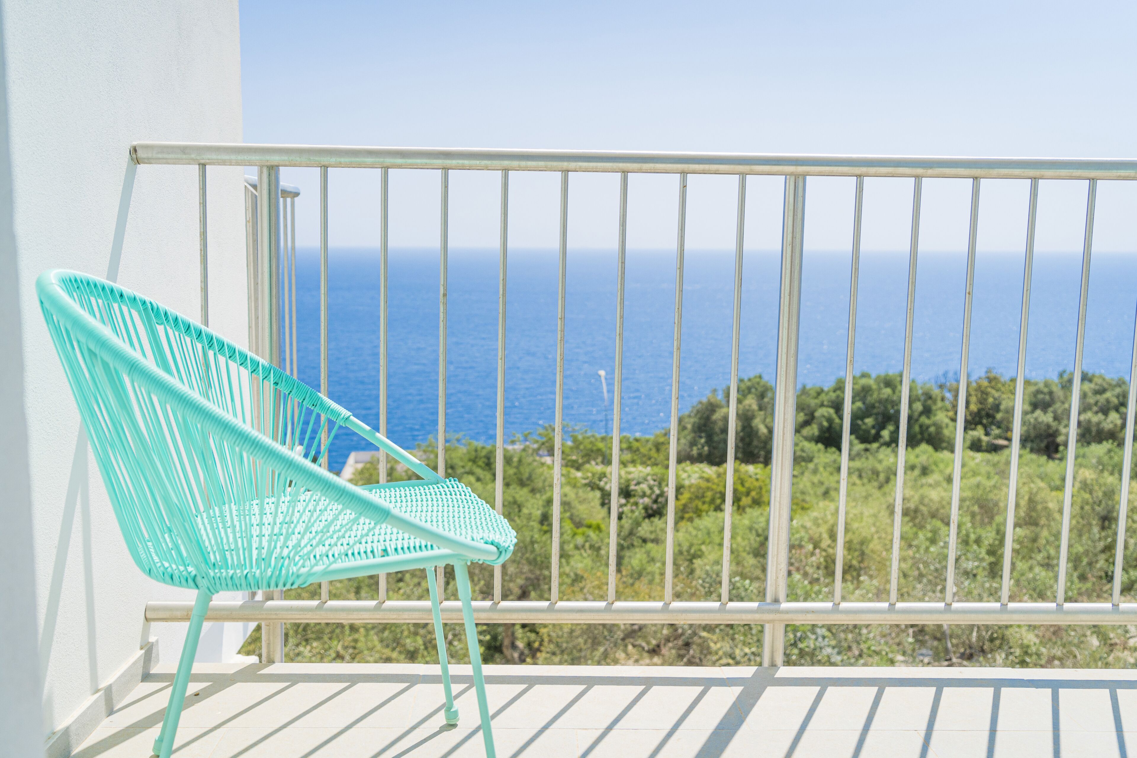 Ein türkisfarbener Stuhl auf einem Balkon mit weißer Reling, mit Blick auf eine Küstenlandschaft mit viel Grün und dem Meer.