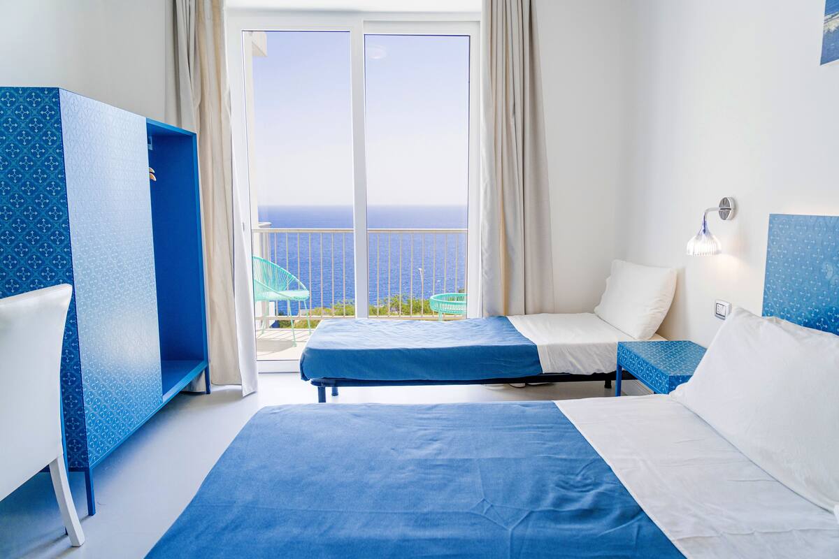 A hotel room with two beds, a desk, and a view of the sea.