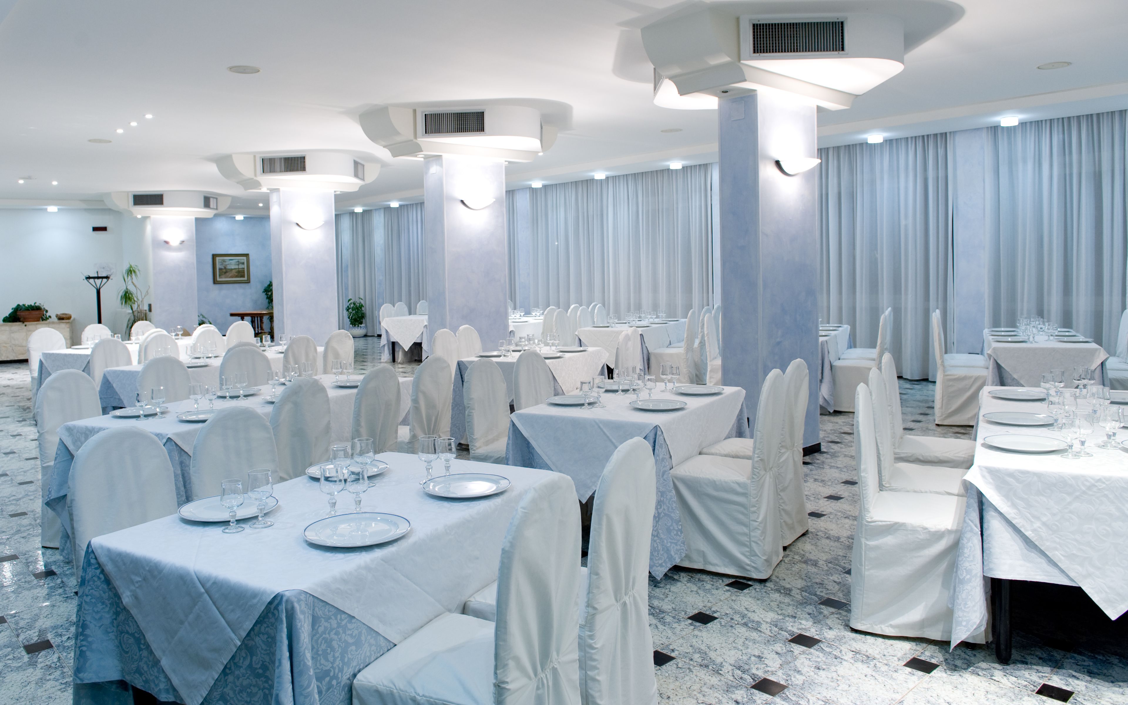 A banquet hall with multiple tables set for dining, featuring white tablecloths and chairs, and a marble floor.