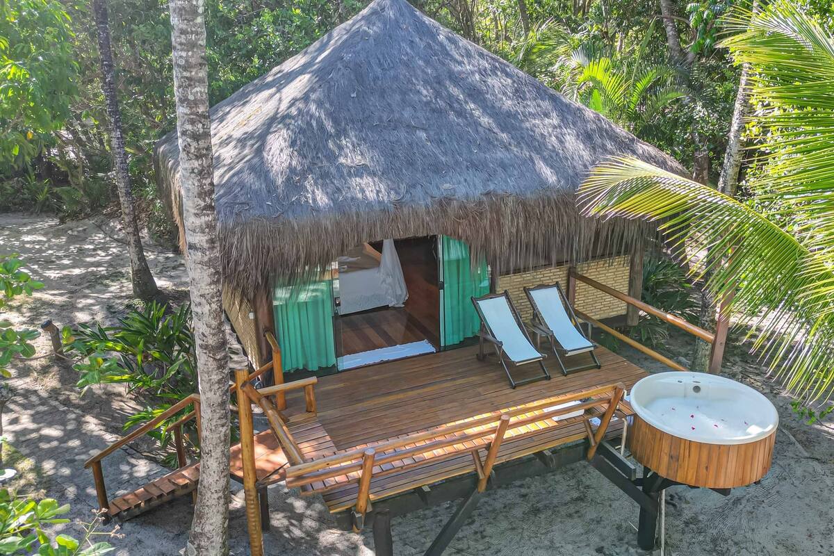 A thatched-roof hut with a wooden deck, two lounge chairs, a small wooden table, and a hot tub.