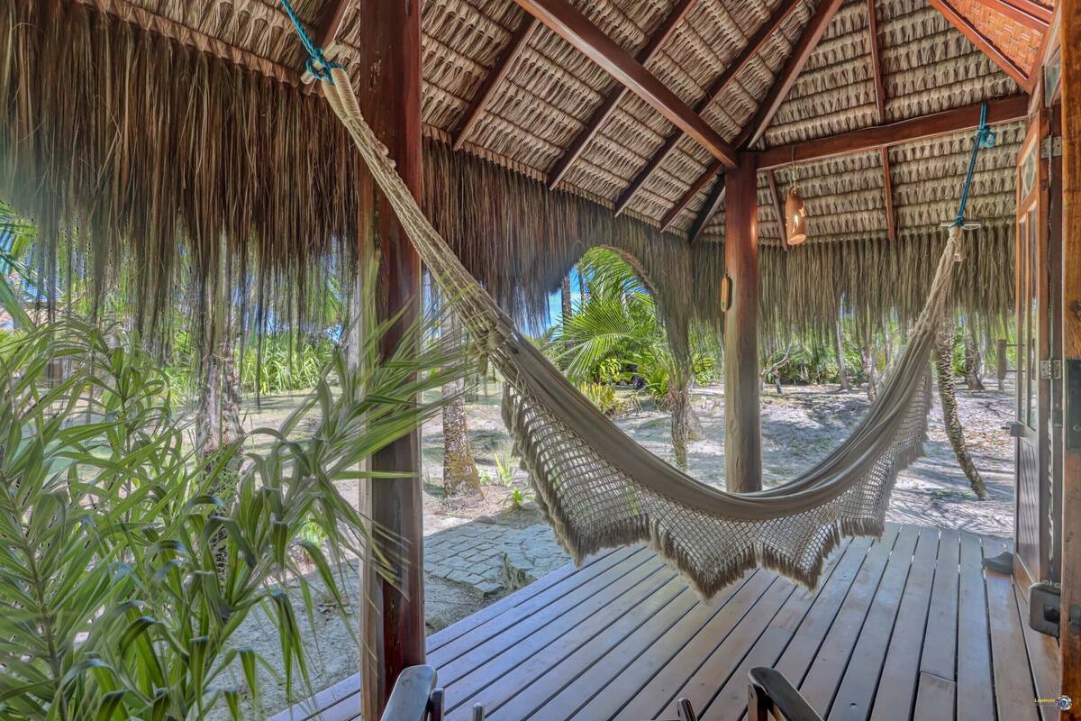 A hammock under a thatched roof structure, wooden deck, and tropical plants.