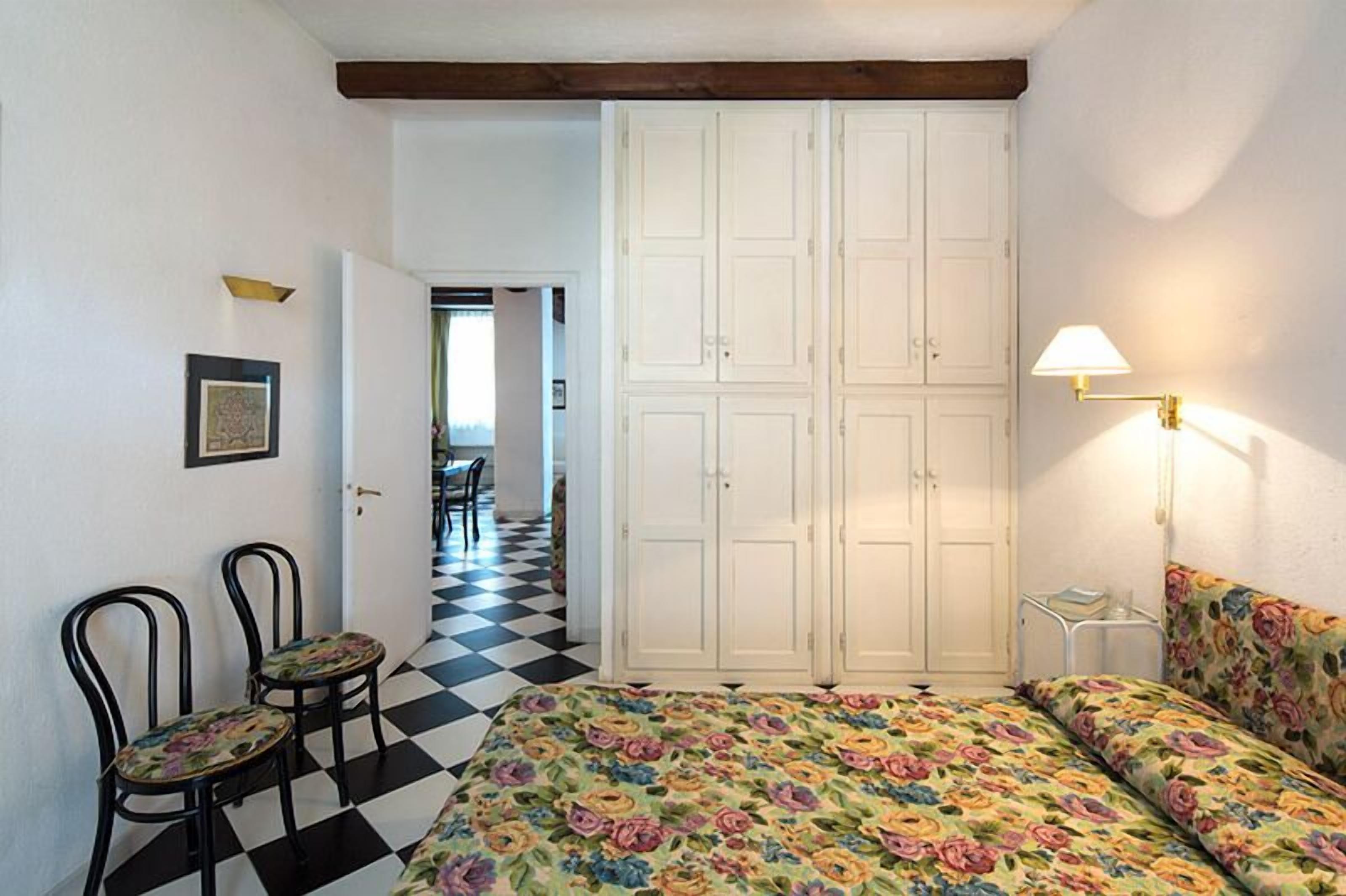 A bedroom with a checkered floor, a bed with floral bedding, two chairs, a wardrobe, and a lamp.