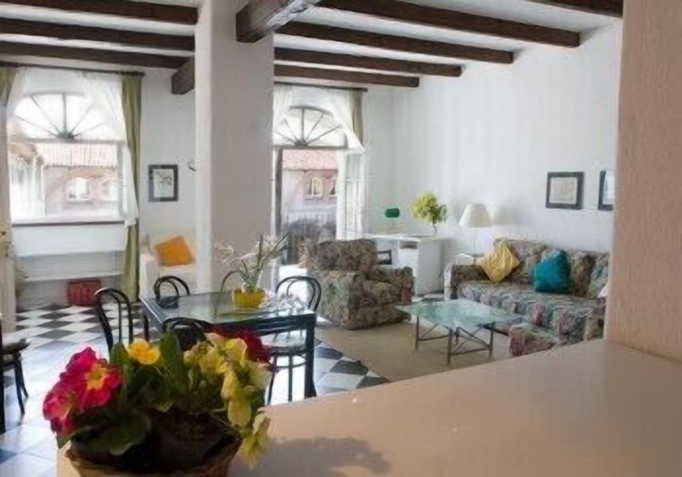 Un salon avec un sol à carreaux, un canapé, une table basse et une table à manger avec des chaises. Il y a des fenêtres avec des rideaux, une lampe sur la table et une plante sur le sol.