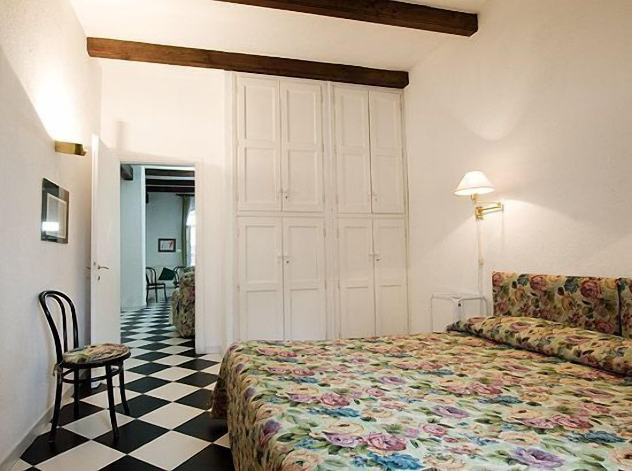 A bedroom with a checkered floor, a bed with a floral bedspread, a chair, a lamp, and a wardrobe.