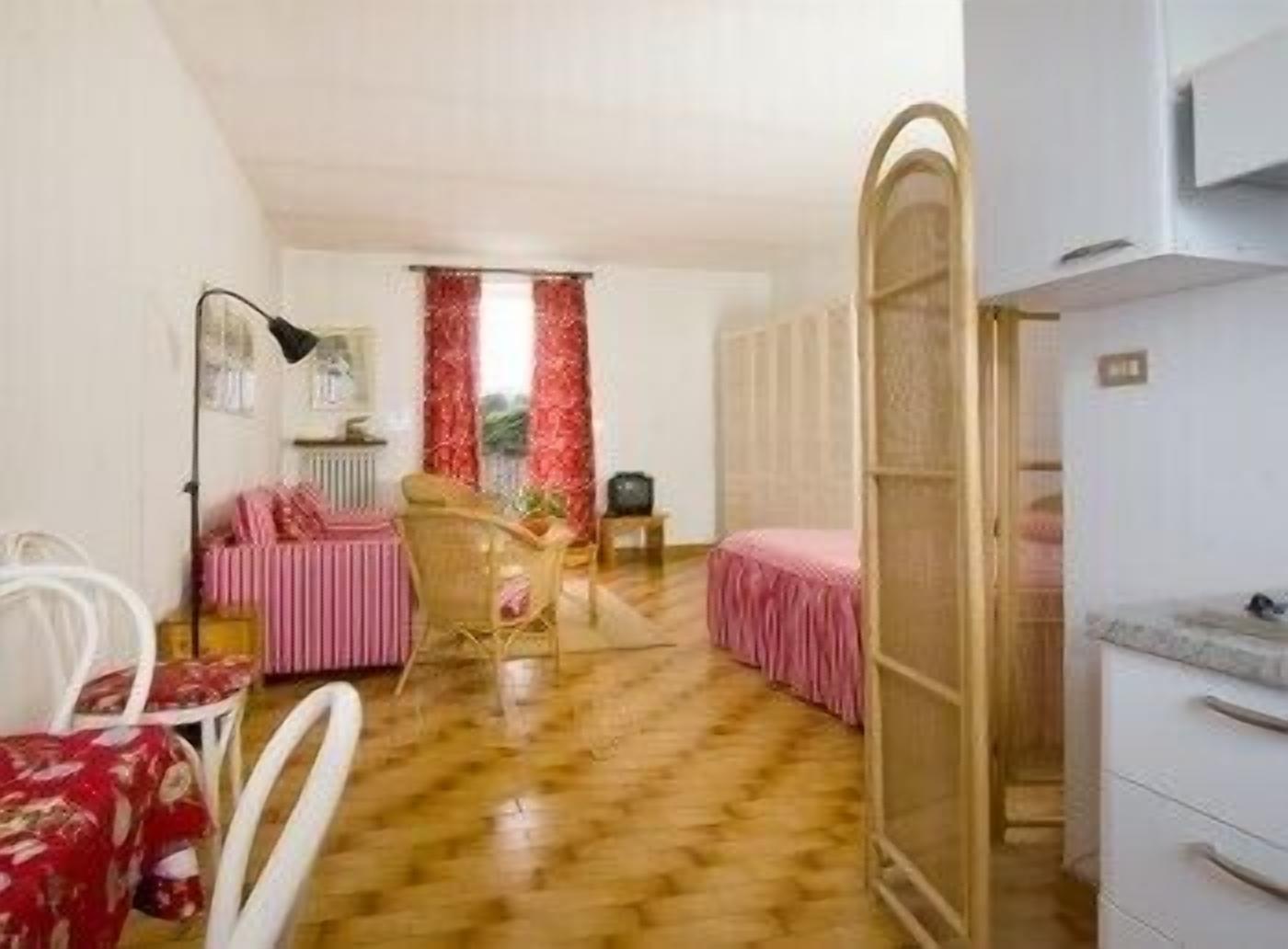 A room with a wooden floor, a bed with a pink bedspread, a chair, a desk with a lamp, and a wardrobe.