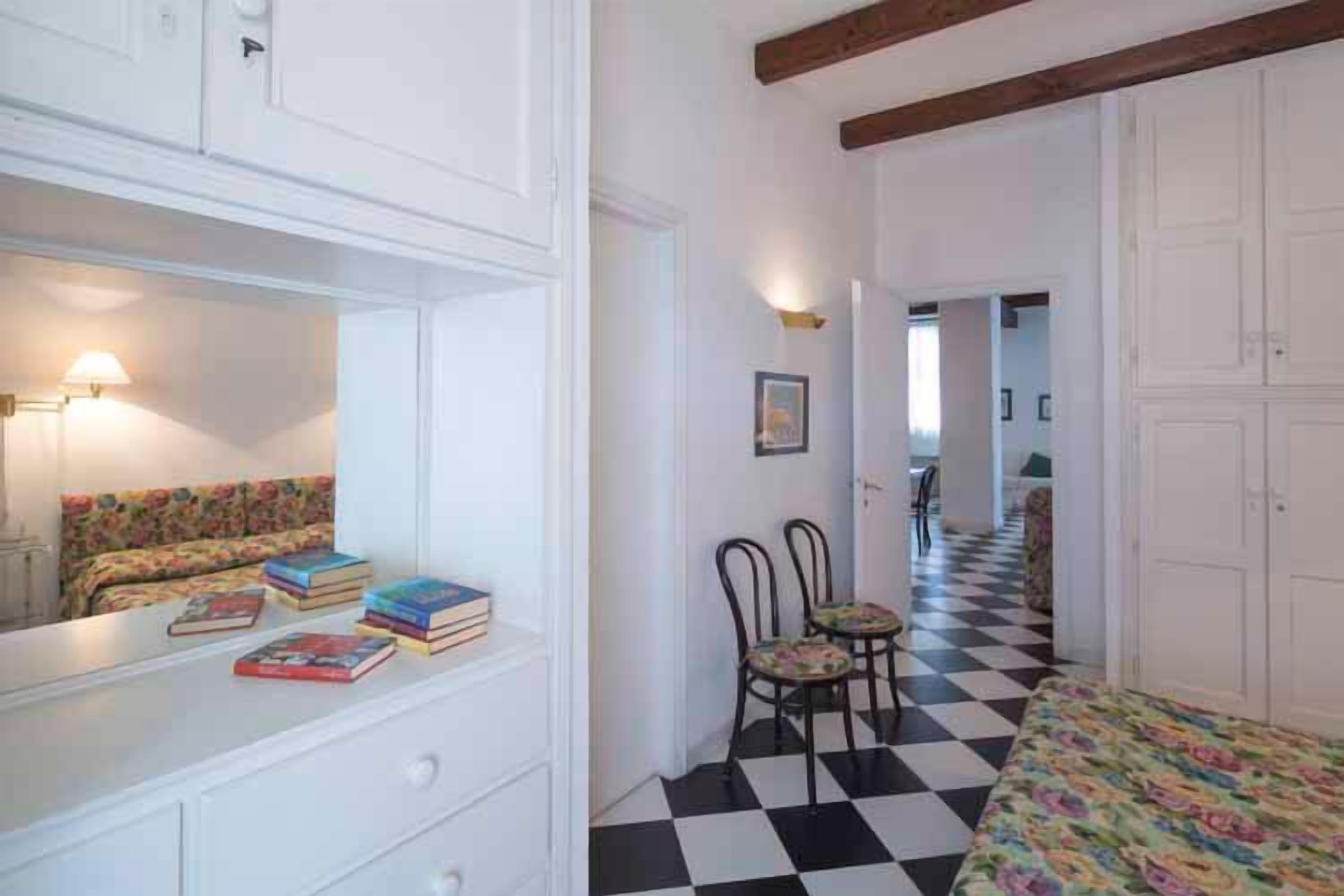Un dormitorio con cama, cómoda, espejo y silla. El piso es de cuadros y hay una puerta que da a otra habitación.