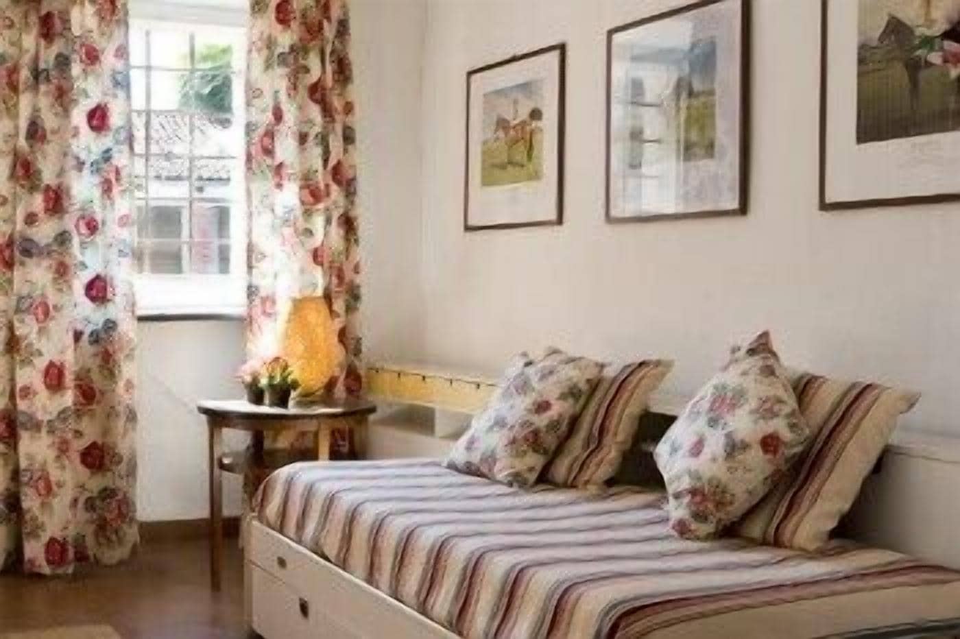 A bedroom with a bed, floral curtains, a small table with a lamp, and two framed pictures on the wall.
