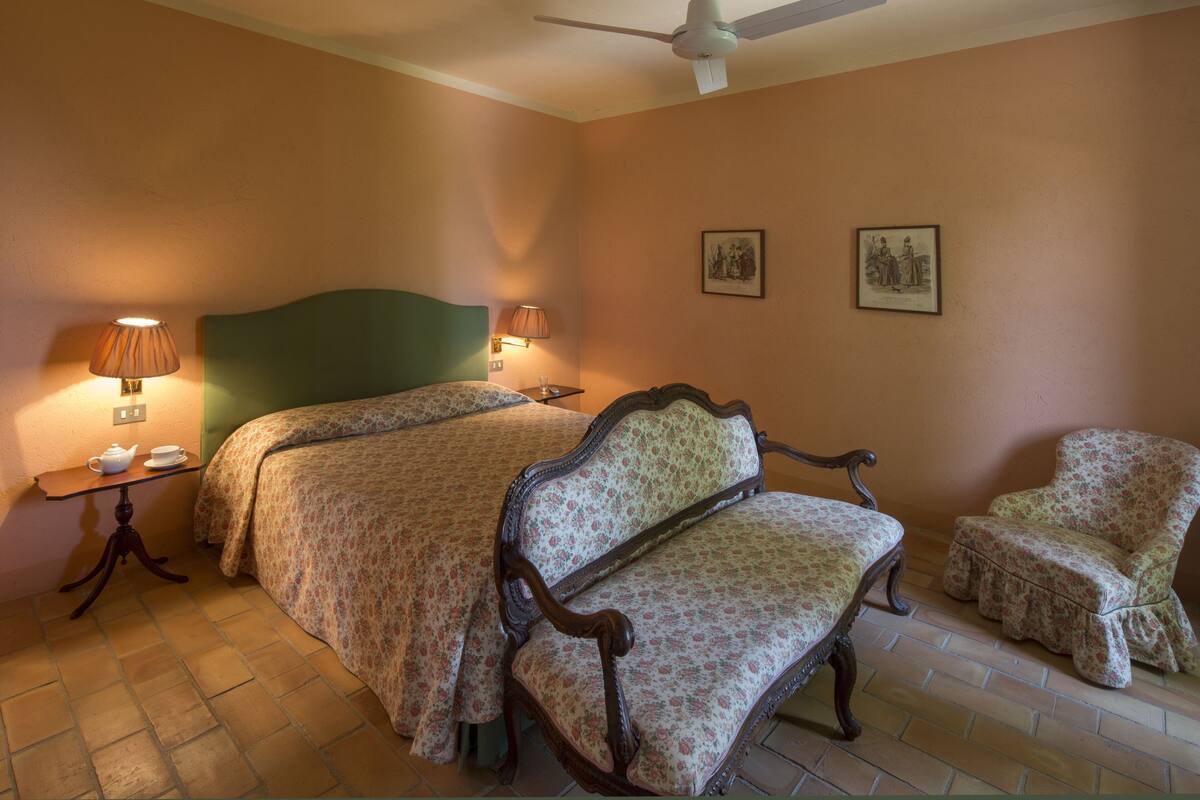 A hotel room with two beds, a chair, a table with a lamp, and two wall-mounted pictures.