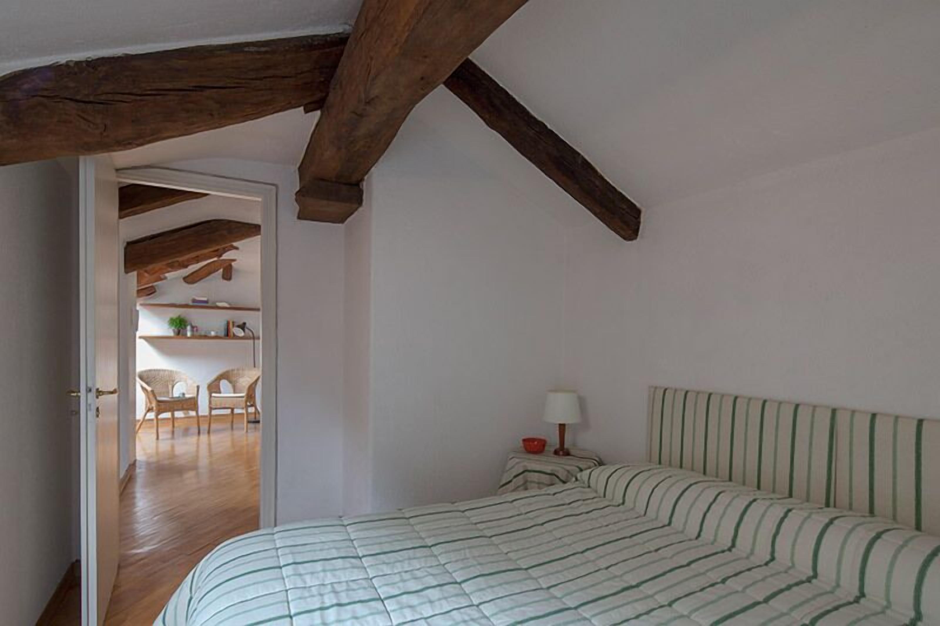 A bedroom with a bed, a nightstand, a lamp, and a wooden ceiling beam.
