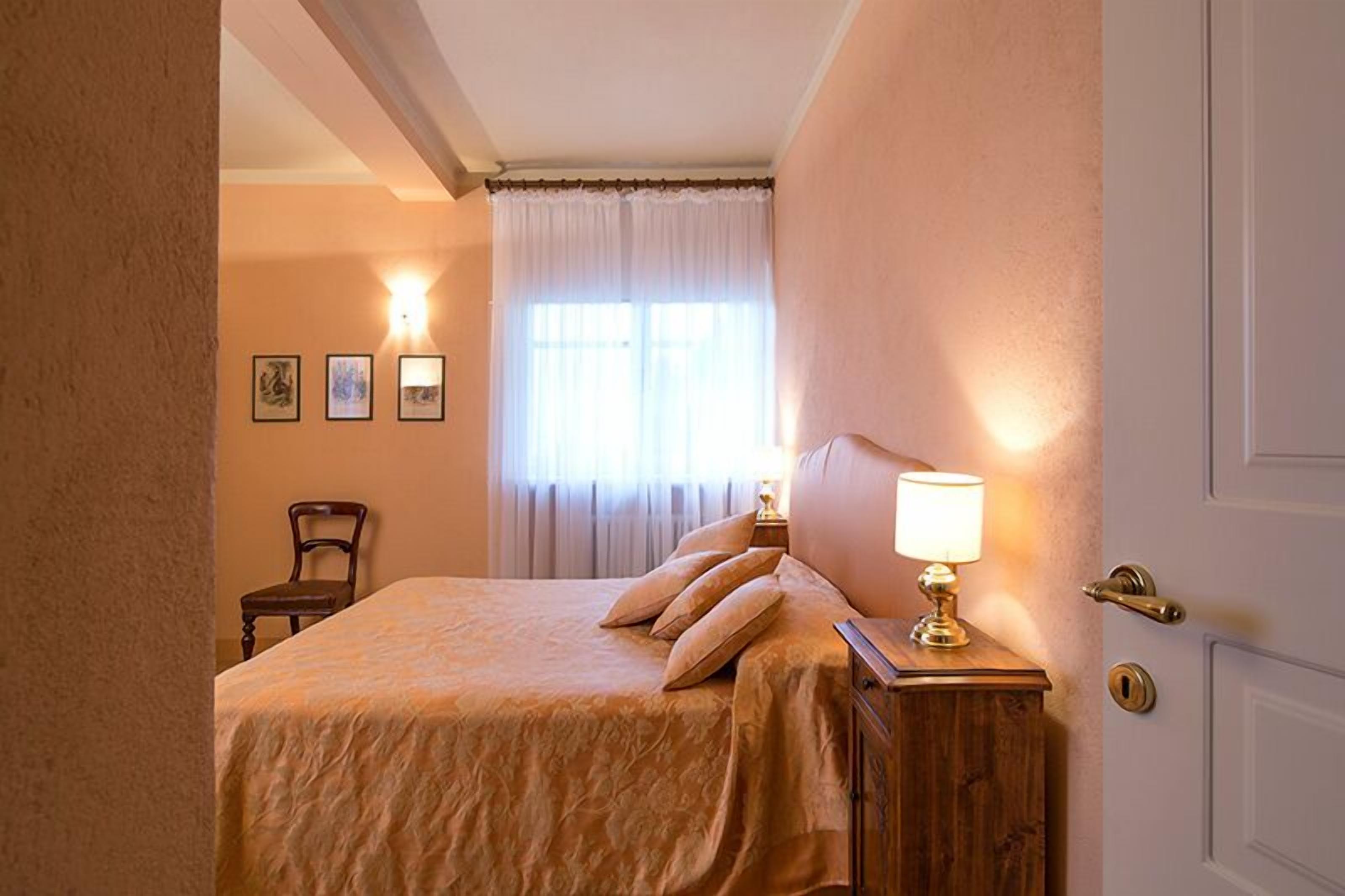 A bedroom with a bed, a nightstand with a lamp, a chair, and a window with curtains.