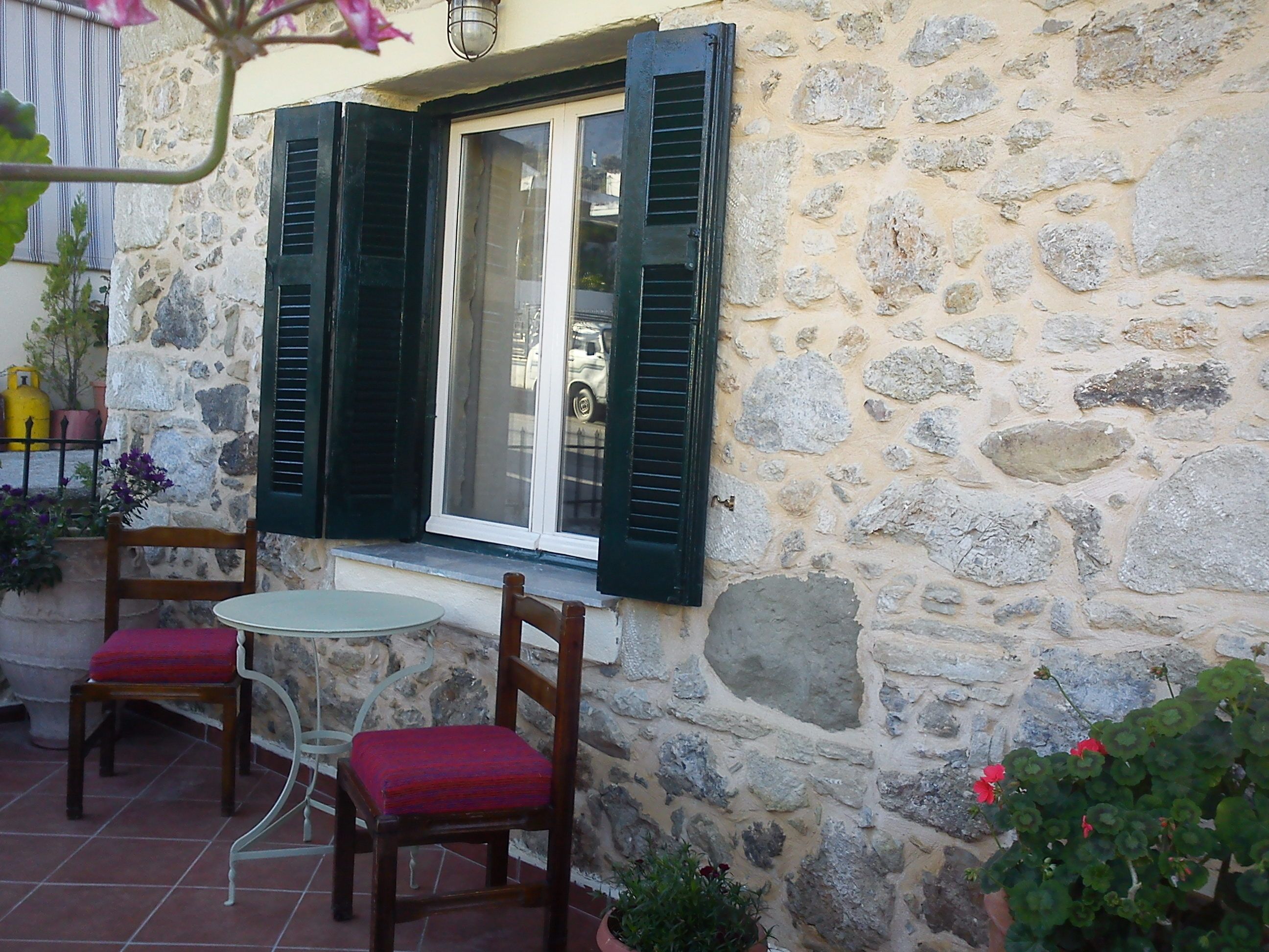A stone building with a window and shutters, a small table, two chairs with red cushions, and potted plants.