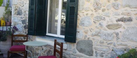 A stone building with a window and shutters, a small table, two chairs with red cushions, and potted plants.
