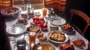 Une table à manger dressée avec différents plats, notamment des tartes et un saladier de fruits, près d’une fenêtre ornée de rideaux.