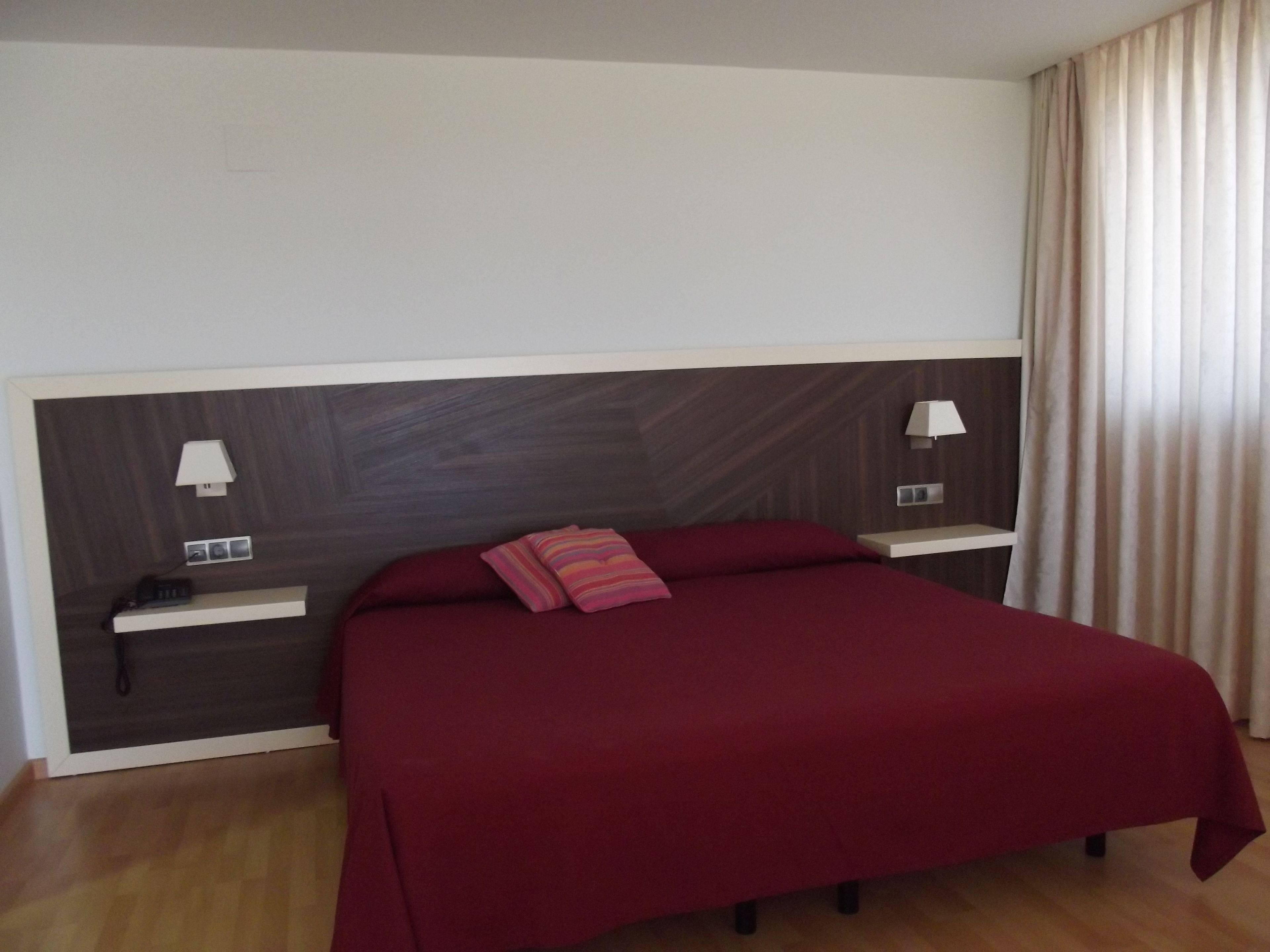 A hotel room with a large bed, two bedside tables with lamps, a power outlet, and a window with curtains.
