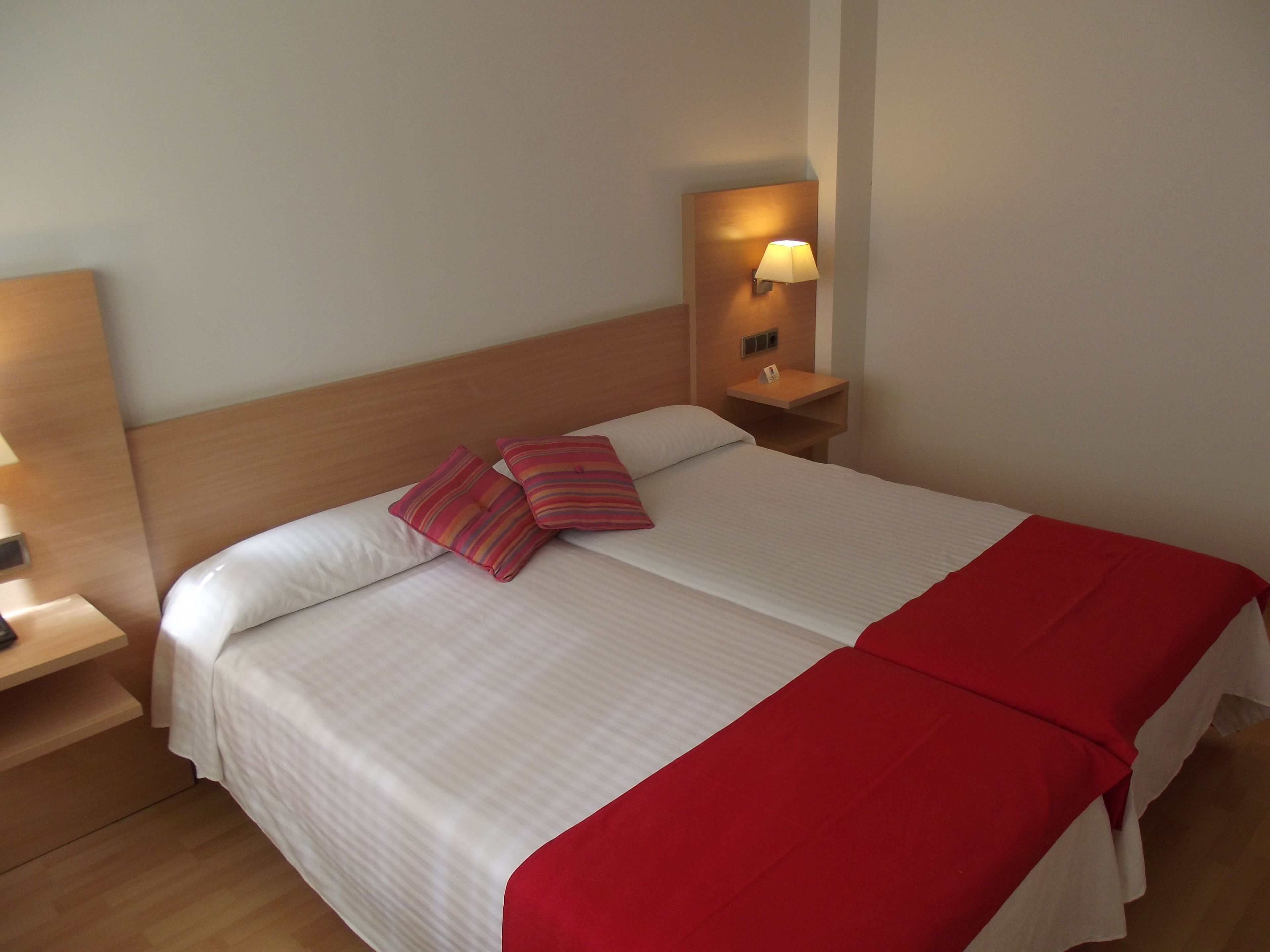 Una cama bien tendida con sábanas blancas y dos almohadas a rayas, un cabecero de madera, una mesita de noche con una lámpara y una manta roja.
