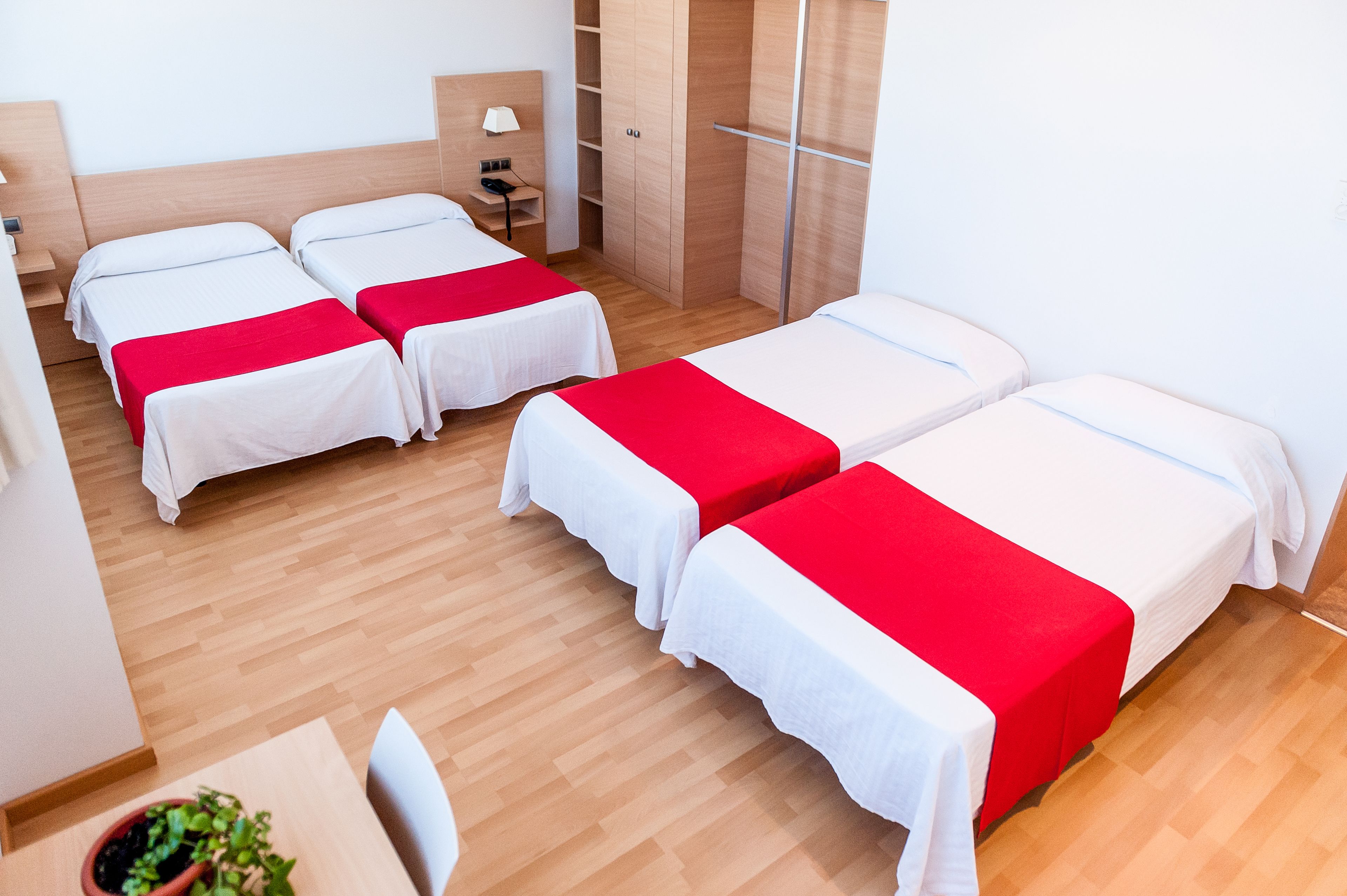 A hotel room with four single beds, wooden flooring, a desk with a chair, a small table with a potted plant, and a wardrobe.