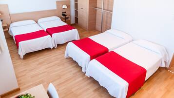 A hotel room with four single beds, wooden flooring, a desk with a chair, a small table with a potted plant, and a wardrobe.
