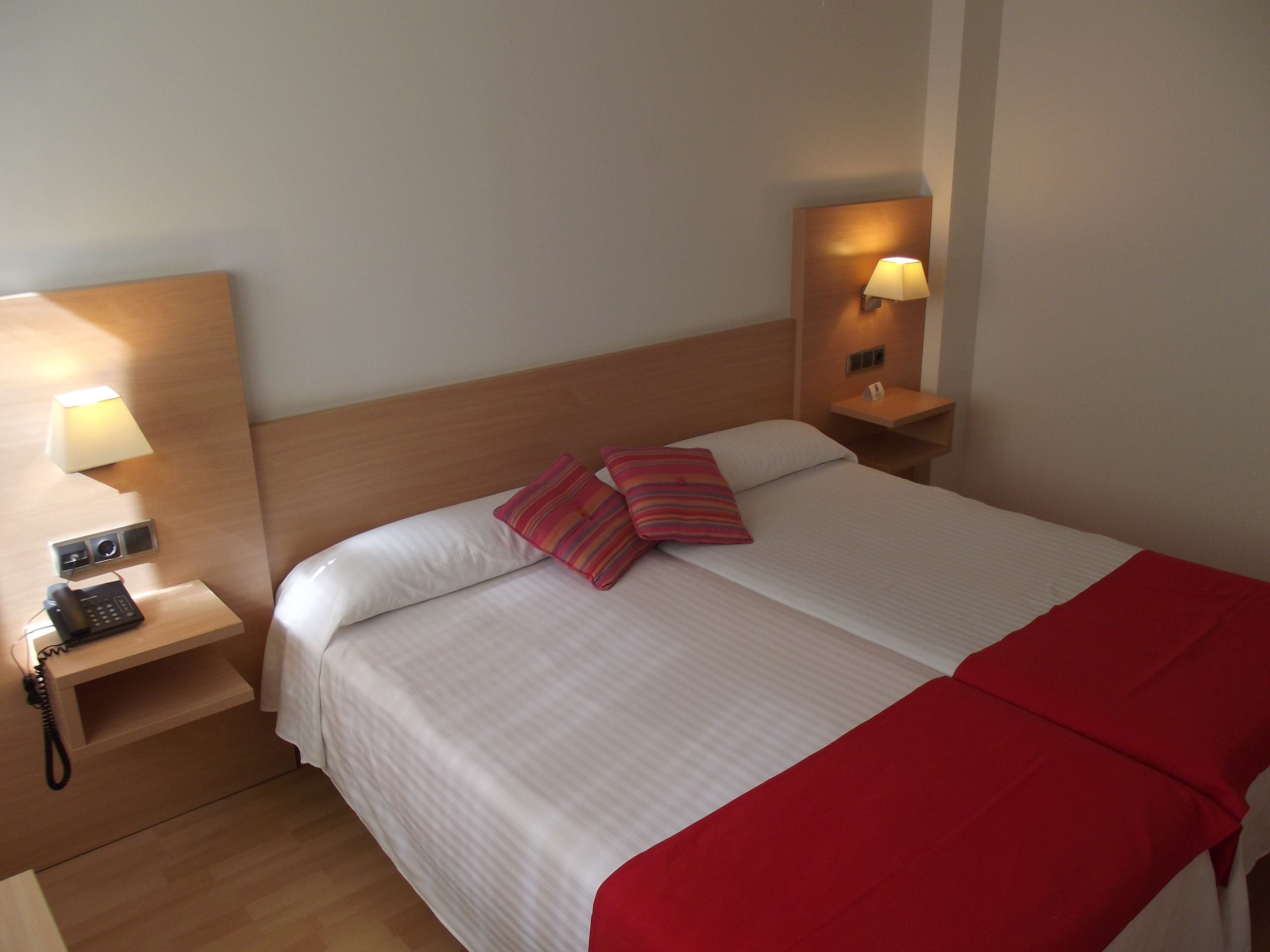 Una camera d'albergo con un letto rifatto, un telefono sul comodino e due lampade a parete.