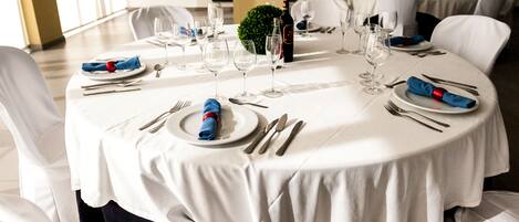A round dining table set with white tablecloths, silverware, and wine glasses, surrounded by white chairs.