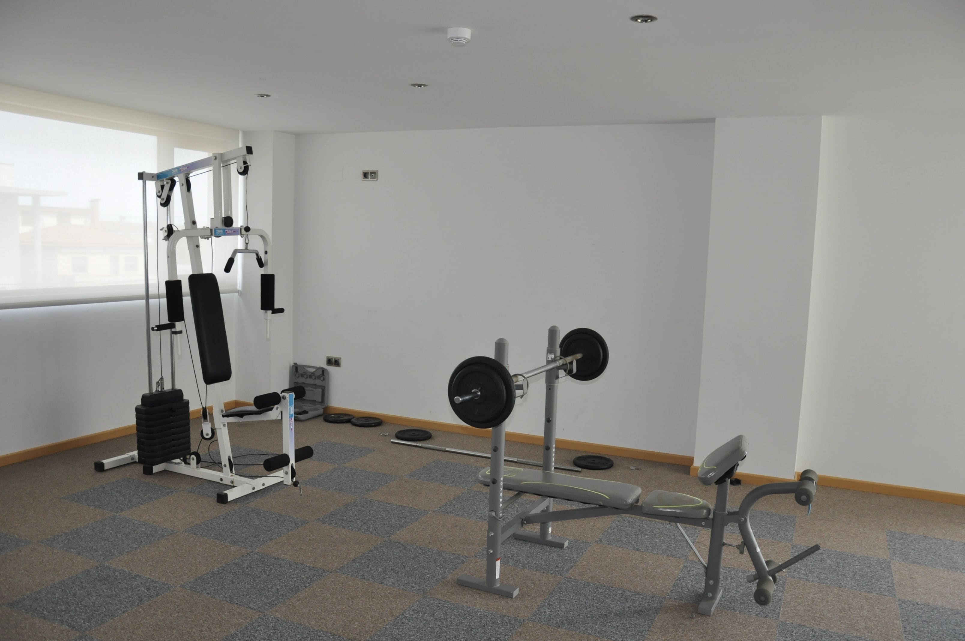 Un gimnasio con equipo de levantamiento de pesas, una banca y una ventana que ofrece vistas a edificios.