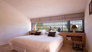 A bedroom with a large bed, a window with a metal grill, a bedside table with a lamp, and a small wooden stool. - Dar Maktoub (Inezgane)