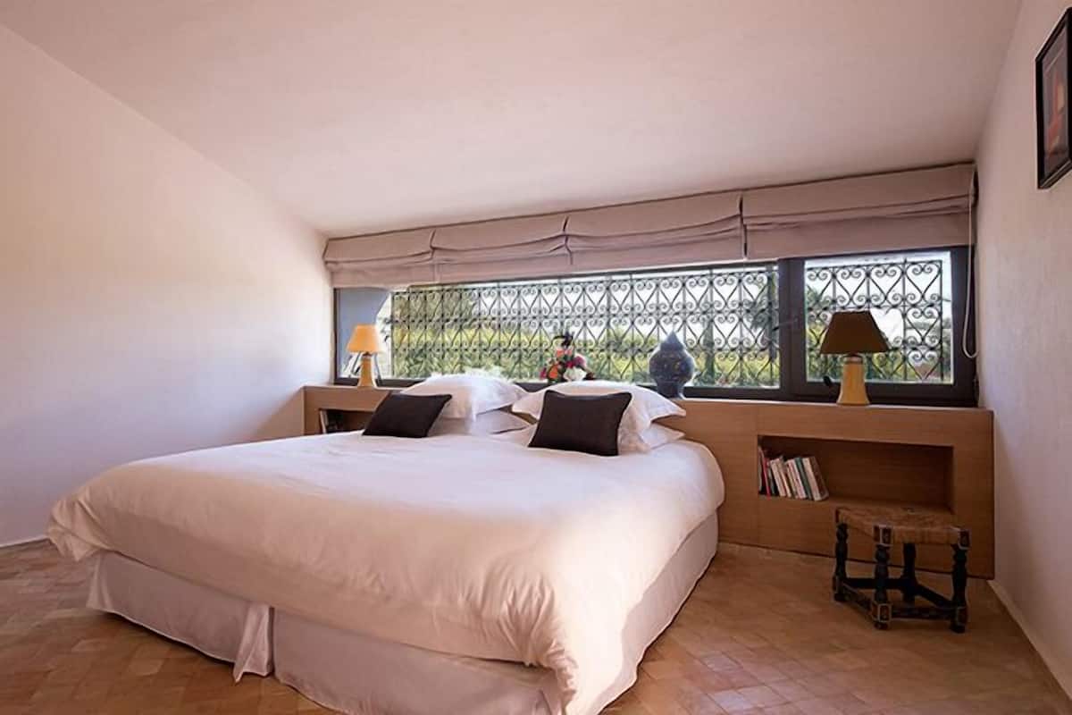 A bedroom with a large bed, a window with a metal grill, a bedside table with a lamp, and a small wooden stool.