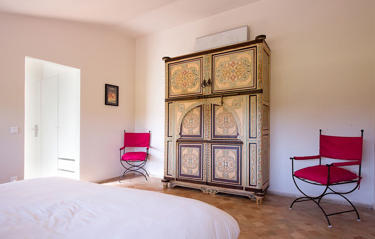 Un dormitorio con una cama grande, dos sillas rojas, un aparador decorativo y una puerta que da a otra habitación.