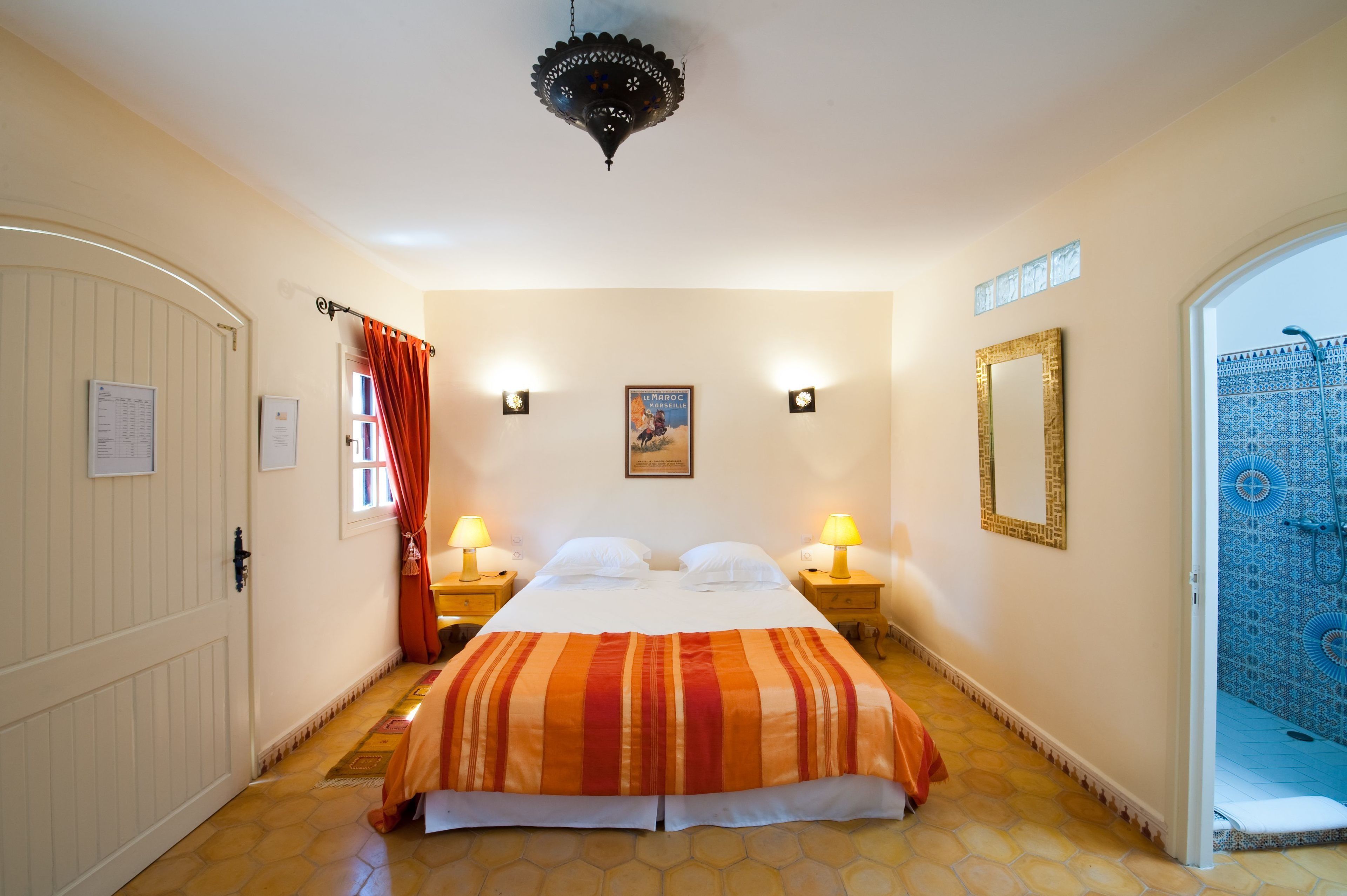 Habitació d'hotel amb un llit, dues làmpades de nit, un mirall decoratiu, una porta amb cortina i vista a una zona amb rajoles blaves.