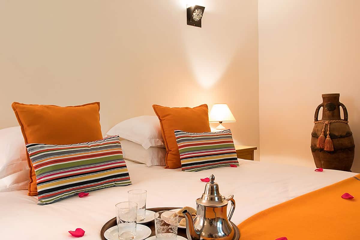 A hotel room with a neatly made bed, orange bedding, a tray with a coffee pot, glasses, and a teapot.